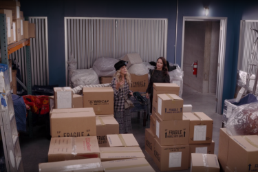 A screenshot from episode 6 of season one of the series "And Just Like That..." featuring the characters Carrie Bradshaw and Charlotte York in Carrie's warehouse, surrounded by carboard boxes